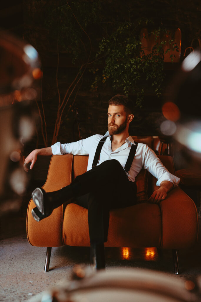 Austin Lowe in a stylish seated pose wearing suspenders and a white shirt, photographed in a warm, moody setting