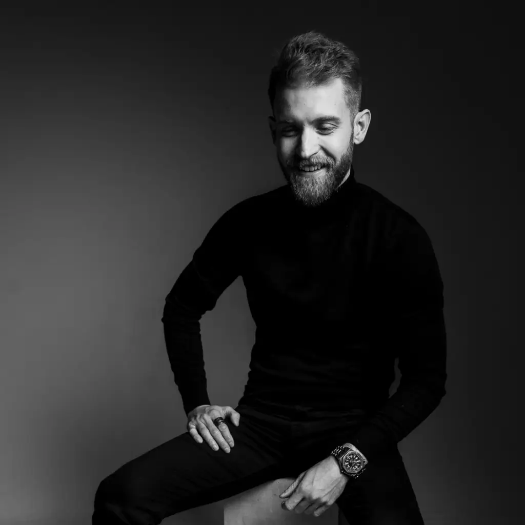 Black and white portrait of a man sitting on a stool, smiling, wearing a dark outfit and a watch in a minimalist studio setting